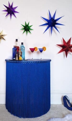 Blue fringed console table with bottles and cocktail glasses against a white wall with brightly coloured stars