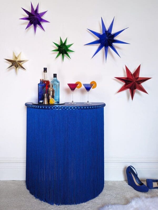 Blue fringed console table with bottles and cocktail glasses against a white wall with brightly coloured stars