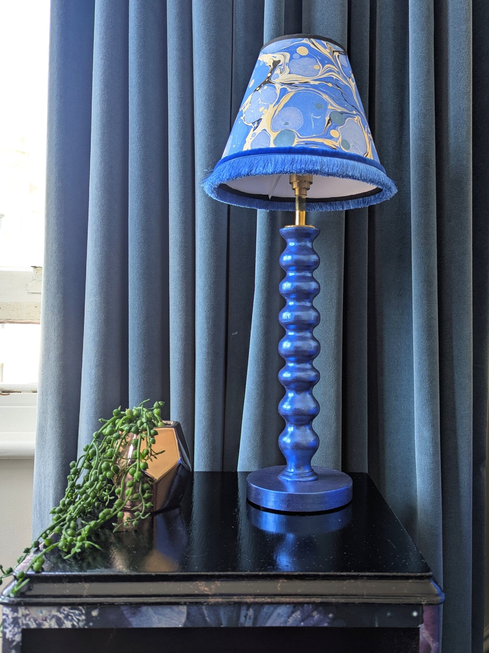 Blue gilded cordless lamp wit blue marbled lampshade on black cabinet in front of blue velvet curtain