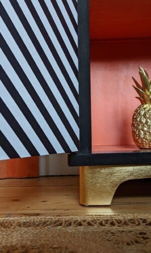 Close up of gold leaf plinth of black and white striped cabinet with door open to show orange interior
