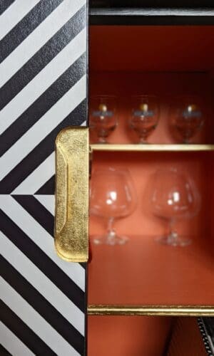 Close up view showing gold leaf recessed handle of black and white striped cabinet, orange interior and gold shelf detail