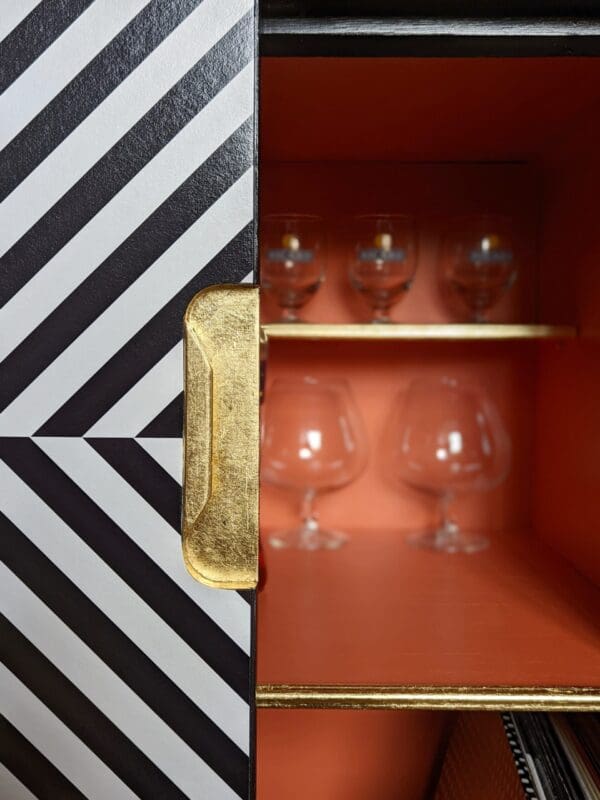 Close up view showing gold leaf recessed handle of black and white striped cabinet, orange interior and gold shelf detail