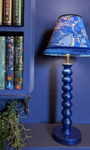 blue metallic bobbin style table lamp with blue marbled lampshade against a blue wall with books