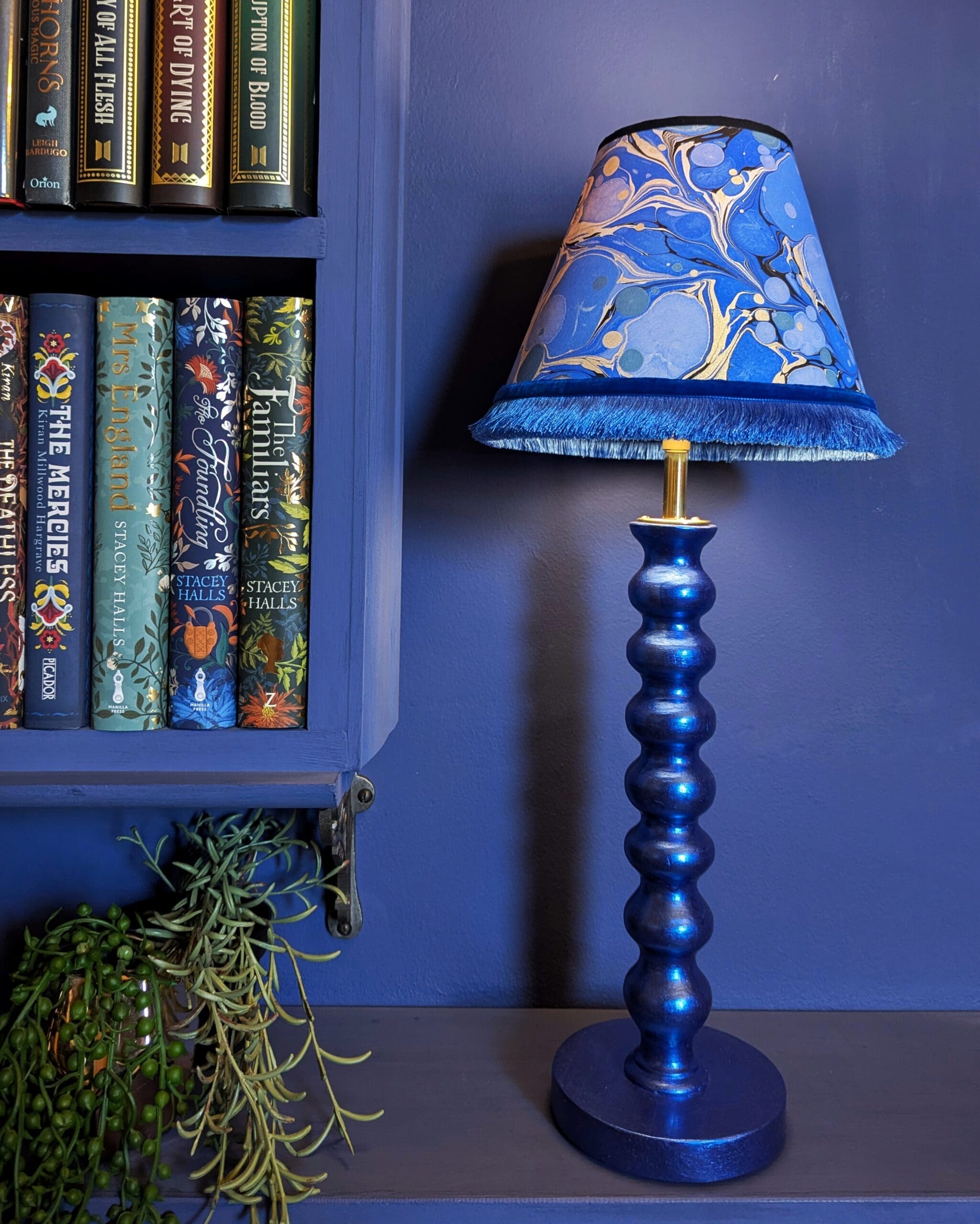blue metallic bobbin style table lamp with blue marbled lampshade against a blue wall with books