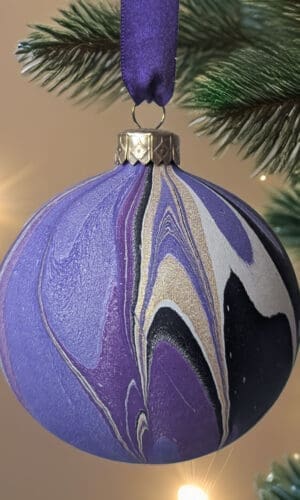 Purple and gold marbled bauble hanging by purple ribbon on a Christmas tree with twinkling lights