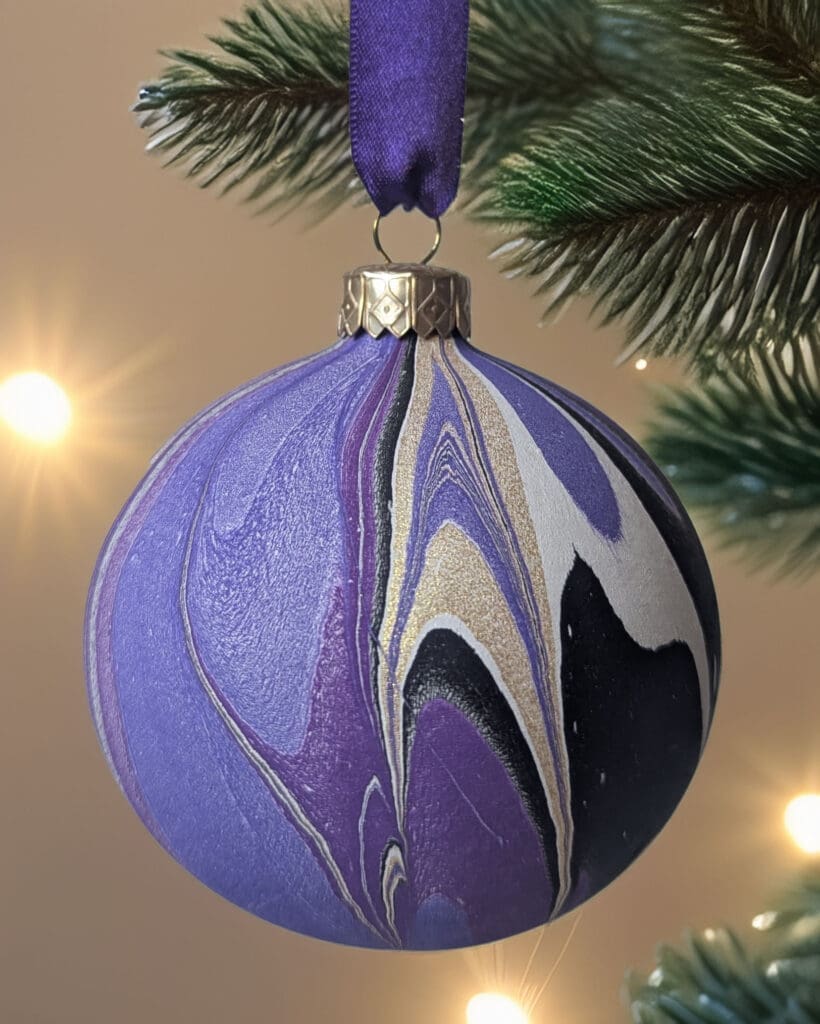 Purple and gold marbled bauble hanging by purple ribbon on a Christmas tree with twinkling lights